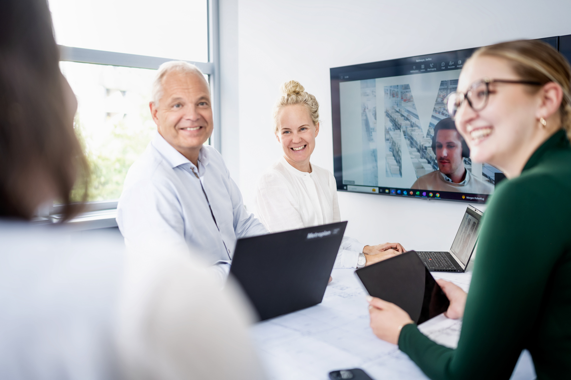 Vier Personen an einem Konferenztisch sitzen mit Laptops und einem Tablet; auf einem Wandbildschirm ist ein weiterer Teilnehmer per Videokonferenz zu sehen; heller Besprechungsraum mit großem Fenster.