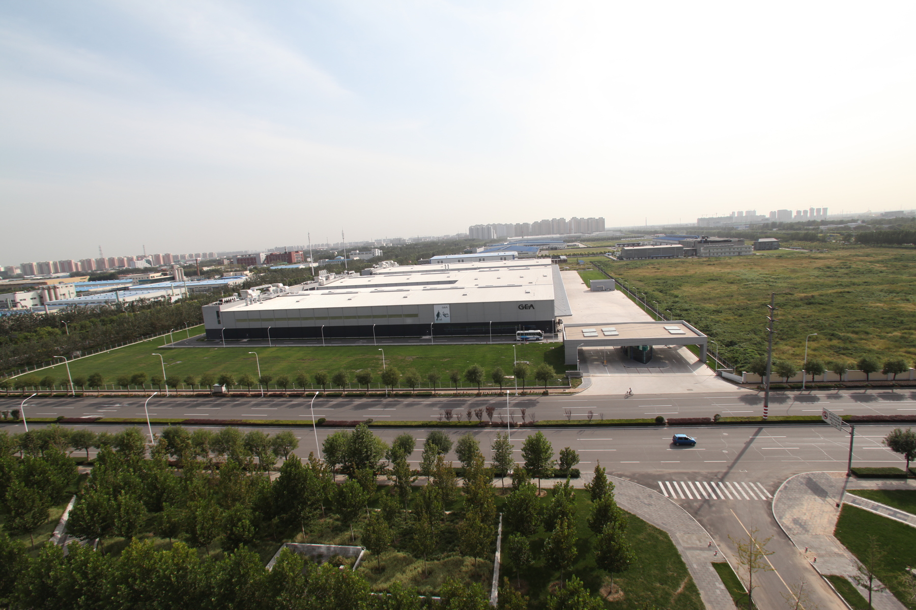 Große Industriehalle mit der Aufschrift "GEA" liegt auf umzäuntem Firmengelände; davor Einfahrt mit Pförtnerhaus. Mehrspurige Straße mit wenigen Fahrzeugen, Rasenflächen und Bäumen; umliegende Fabriken und Wohnblöcke am Horizont.