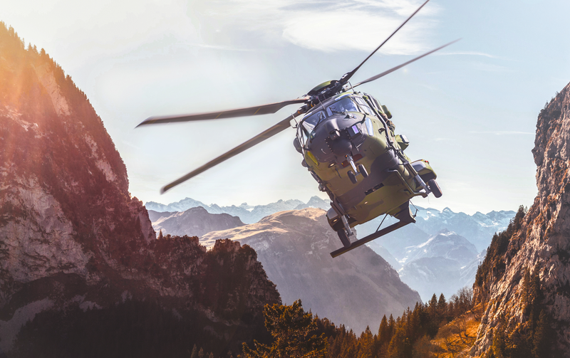 Mehrzweckhubschrauber fliegt schräg aufwärts durch ein enges Gebirgstal, Rotorblätter in Bewegung; umgeben von steilen Felswänden, schneebedeckten Gipfeln und Nadelwald, warmes Sonnenlicht, klare Fernsicht.