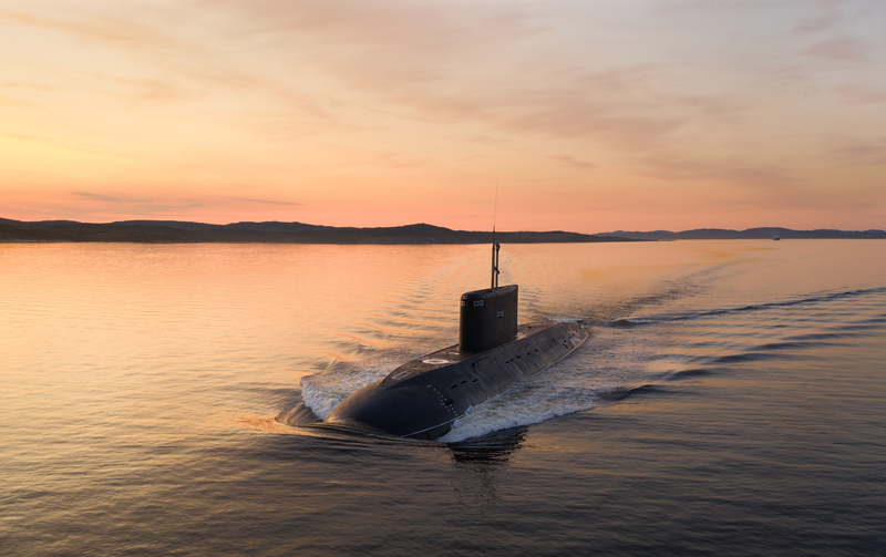Schwarzes U-Boot fährt halb aufgetaucht durch ruhiges Wasser und hinterlässt eine weiße Bugwelle; orangefarbener Sonnenuntergang spiegelt sich, ferne Uferlinien und sanfte Hügel am Horizont.
