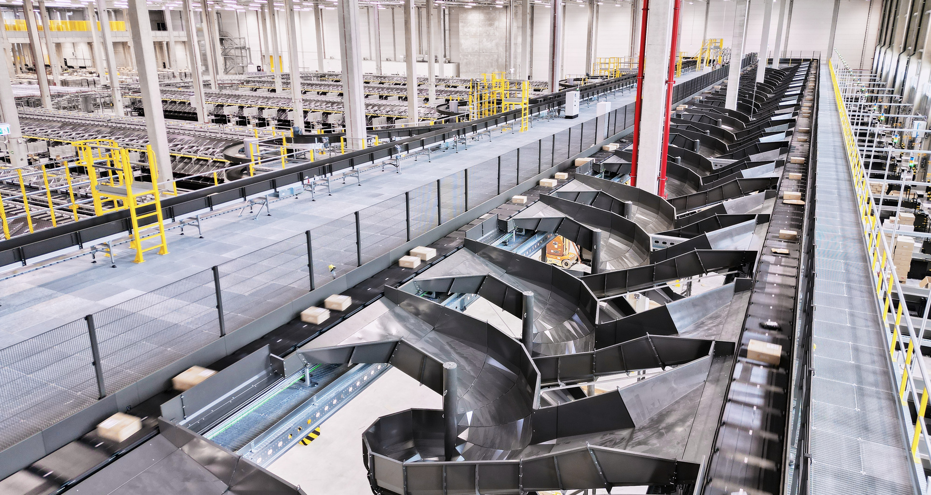 Automatisches Förderbandsystem mit Kartonpaketen bewegt und sortiert Sendungen über Weichen in seitliche Rutschen; große Lagerhalle mit Metallstegen, Geländern, Stützpfeilern, hellen Deckenleuchten und zahlreichen Förderlinien.